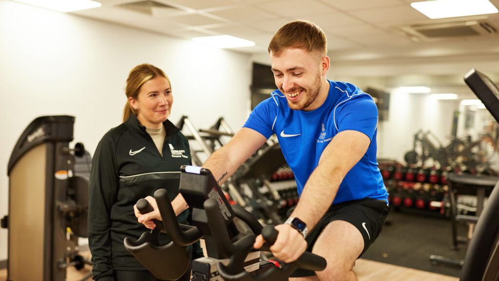 Young cheerful female personal coach helping young male doing cycling workout in gym
