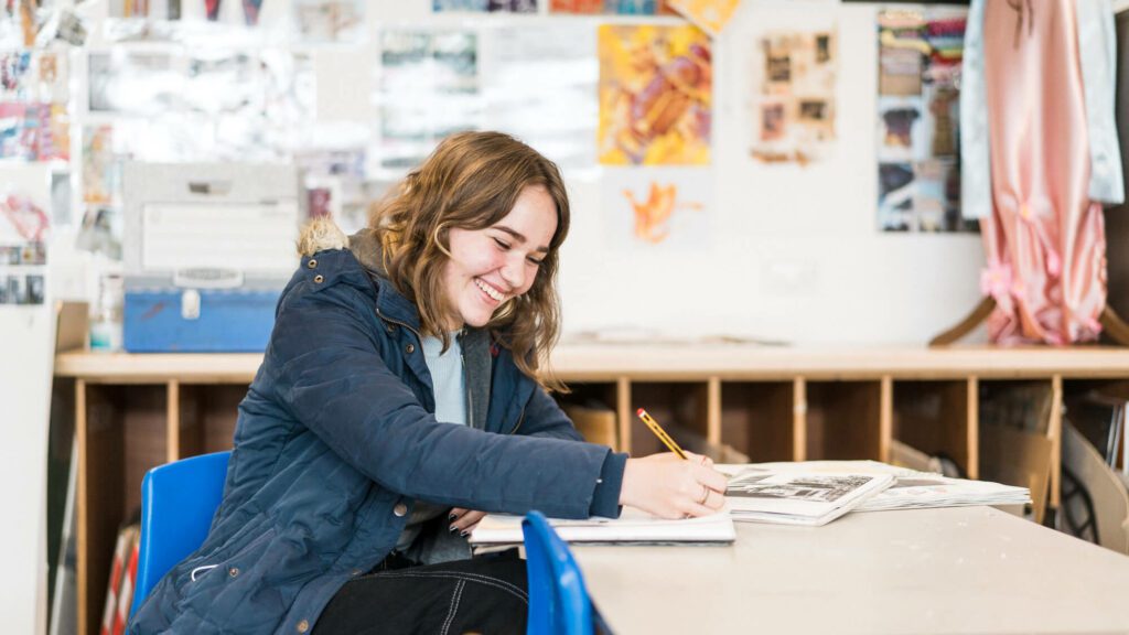 Female student sat at desk writing and smiling