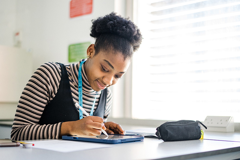 Student wearing blue lanyard sitting in classroom writing on iPad with notes on the table